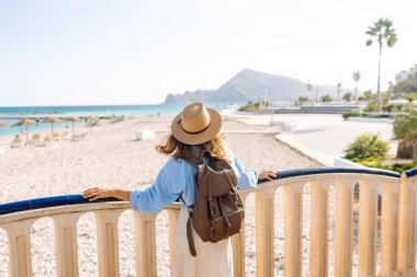 Şapkalı ve sırt çantalı bir gezgin güneşli şehir seti boyunca geziniyor. Arkadan bak. Sırt çantalı güzel bir kadın deniz manzarasını seviyor. Doğa, turizm ve özgürlük kavramsal fikirleri.