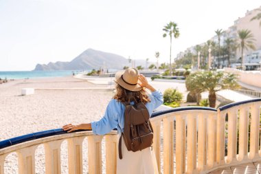 Şapkalı ve sırt çantalı bir gezgin güneşli şehir seti boyunca geziniyor. Arkadan bak. Sırt çantalı güzel bir kadın deniz manzarasını seviyor. Doğa, turizm ve özgürlük kavramsal fikirleri.