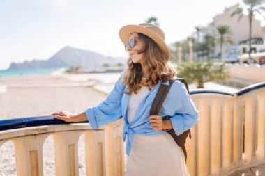 Şapkalı ve sırt çantalı bir gezgin güneşli şehir seti boyunca geziniyor. Arkadan bak. Sırt çantalı güzel bir kadın deniz manzarasını seviyor. Doğa, turizm ve özgürlük kavramsal fikirleri.