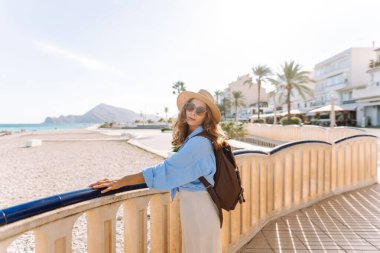 Şapkalı ve sırt çantalı bir gezgin güneşli şehir seti boyunca geziniyor. Arkadan bak. Sırt çantalı güzel bir kadın deniz manzarasını seviyor. Doğa, turizm ve özgürlük kavramsal fikirleri.