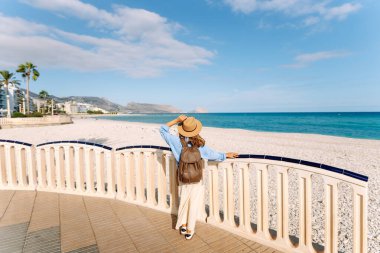 Şapkalı ve sırt çantalı bir gezgin güneşli şehir seti boyunca geziniyor. Arkadan bak. Sırt çantalı güzel bir kadın deniz manzarasını seviyor. Doğa, turizm ve özgürlük kavramsal fikirleri.