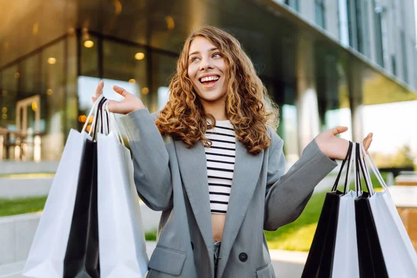 Modern bir binanın önünde alışveriş torbalarıyla şık kıyafetler içinde neşeli bir kadın. Şık bir kadın alışverişten sonra güneşli bir günün tadını çıkarır. Tüketim kavramı, alışveriş.