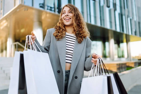 Modern bir binanın önünde alışveriş torbalarıyla şık kıyafetler içinde neşeli bir kadın. Şık bir kadın alışverişten sonra güneşli bir günün tadını çıkarır. Tüketim kavramı, alışveriş.