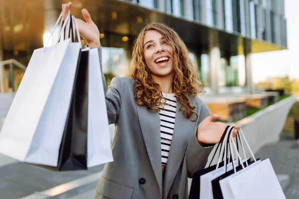 Modern bir binanın önünde alışveriş torbalarıyla şık kıyafetler içinde neşeli bir kadın. Şık bir kadın alışverişten sonra güneşli bir günün tadını çıkarır. Tüketim kavramı, alışveriş.