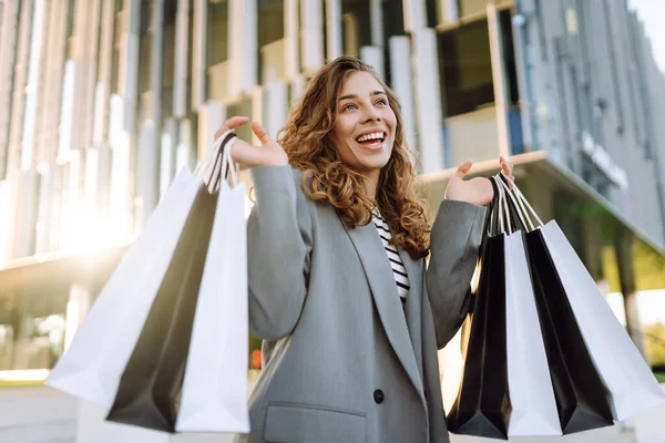 Modern bir binanın önünde alışveriş torbalarıyla şık kıyafetler içinde neşeli bir kadın. Şık bir kadın alışverişten sonra güneşli bir günün tadını çıkarır. Tüketim kavramı, alışveriş.
