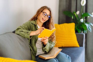 Sarı yastıklı genç bir kadın kanepeye oturur ve sarı bir deftere yazar. Gülümseyen kadın rahat bir odada rahatlar, notlar alır, yalnız vakit geçirir. Konfor, eğitim, rahatlama kavramları.