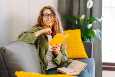 Sarı yastıklı genç bir kadın kanepeye oturur ve sarı bir deftere yazar. Gülümseyen kadın rahat bir odada rahatlar, notlar alır, yalnız vakit geçirir. Konfor, eğitim, rahatlama kavramları.