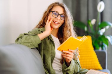 Sarı yastıklı genç bir kadın kanepeye oturur ve sarı bir deftere yazar. Gülümseyen kadın rahat bir odada rahatlar, notlar alır, yalnız vakit geçirir. Konfor, eğitim, rahatlama kavramları.