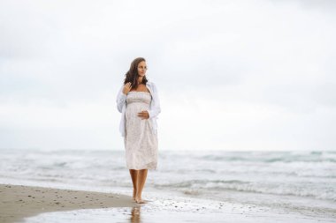 Genç hamile bir kadın sahilde yürüyor, deniz manzarasının tadını çıkarıyor. Elbise giyen hamile kadın, temiz havayı hisseder. Beklenti, rahatlama ve doğa kavramları.