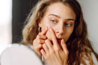 A close-up of a young woman touching her face and looking into a small mirror on a sunny day. A beautiful woman examines a breakout on her face in a cozy room. Concepts: cosmetology, acne, skincare.