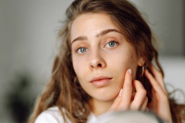 Yüzüne dokunan ve güneşli bir günde küçük bir aynaya bakan genç bir kadının yakın çekimi. Güzel bir kadın, sıcak bir odada yüzündeki patlağı inceliyor. Kozmetik, sivilce, cilt bakımı.