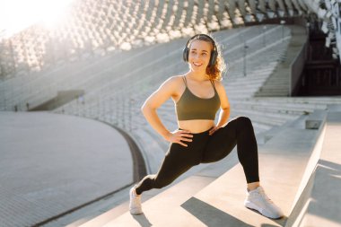 Dışarıda kablosuz kulaklıkla egzersiz yapan genç bir kadın. Atletik bir kadın güneşli bir günde egzersiz yapar, antrenman yapar ve müzik dinler. Spor ve teknoloji kavramları.