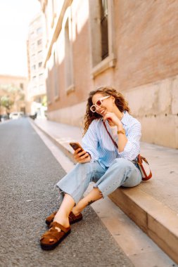 Kıvırcık saçlı, elinde telefon olan, güneşli şehir sokaklarında gezen mutlu bir kadının portresi. Güneş gözlüklü güzel bir turist, elinde telefonla güneşli bir günün ve mimarinin tadını çıkarıyor. Turizm konsepti, şehir.