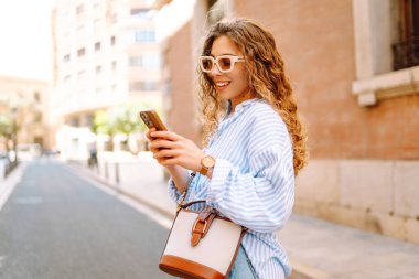 Kıvırcık saçlı, elinde telefon olan, güneşli şehir sokaklarında gezen mutlu bir kadının portresi. Güneş gözlüklü güzel bir turist, elinde telefonla güneşli bir günün ve mimarinin tadını çıkarıyor. Turizm konsepti, şehir.