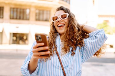 Kıvırcık saçlı, elinde telefon olan, güneşli şehir sokaklarında gezen mutlu bir kadının portresi. Güneş gözlüklü güzel bir turist, elinde telefonla güneşli bir günün ve mimarinin tadını çıkarıyor. Turizm konsepti, şehir.