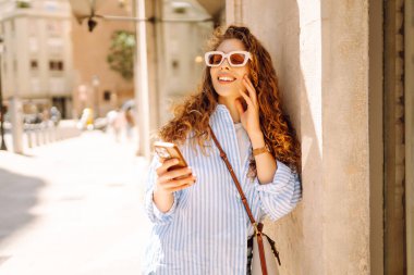 Kıvırcık saçlı, elinde telefon olan, güneşli şehir sokaklarında gezen mutlu bir kadının portresi. Güneş gözlüklü güzel bir turist, elinde telefonla güneşli bir günün ve mimarinin tadını çıkarıyor. Turizm konsepti, şehir.