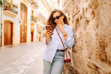Kıvırcık saçlı, elinde telefon olan, güneşli şehir sokaklarında gezen mutlu bir kadının portresi. Güneş gözlüklü güzel bir turist, elinde telefonla güneşli bir günün ve mimarinin tadını çıkarıyor. Turizm konsepti, şehir.