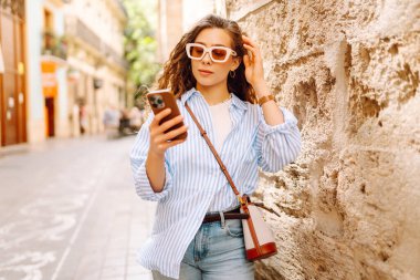 Portrait of a happy woman with curly hair, holding phone, strolling along sunny city streets. Beautiful tourist in sunglasses, holding phone enjoys sunny day and architecture. Tourism concept, urban.