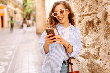 Portrait of a happy woman with curly hair, holding phone, strolling along sunny city streets. Beautiful tourist in sunglasses, holding phone enjoys sunny day and architecture. Tourism concept, urban.