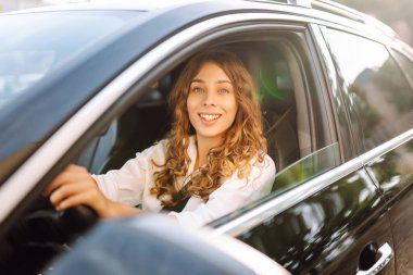 Kıvırcık saçlı genç bir kadın güneşin altında bir arabanın direksiyonunda oturur. Bir kadın sürücü güneşli bir günün tadını çıkarır. Taksi şöförü. Ulaşım kavramı, boş zaman.