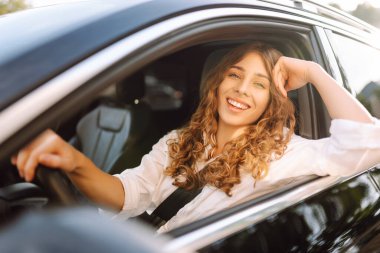 Kıvırcık saçlı genç bir kadın güneşin altında bir arabanın direksiyonunda oturur. Bir kadın sürücü güneşli bir günün tadını çıkarır. Taksi şöförü. Ulaşım kavramı, boş zaman.