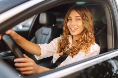 Kıvırcık saçlı genç bir kadın güneşin altında bir arabanın direksiyonunda oturur. Bir kadın sürücü güneşli bir günün tadını çıkarır. Taksi şöförü. Ulaşım kavramı, boş zaman.