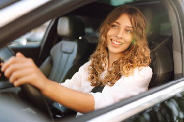 Kıvırcık saçlı genç bir kadın güneşin altında bir arabanın direksiyonunda oturur. Bir kadın sürücü güneşli bir günün tadını çıkarır. Taksi şöförü. Ulaşım kavramı, boş zaman.