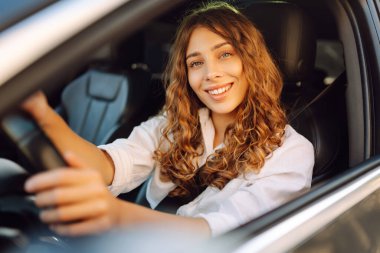 Kıvırcık saçlı genç bir kadın güneşin altında bir arabanın direksiyonunda oturur. Bir kadın sürücü güneşli bir günün tadını çıkarır. Taksi şöförü. Ulaşım kavramı, boş zaman.