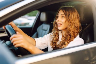 Kıvırcık saçlı genç bir kadın güneşin altında bir arabanın direksiyonunda oturur. Bir kadın sürücü güneşli bir günün tadını çıkarır. Taksi şöförü. Ulaşım kavramı, boş zaman.