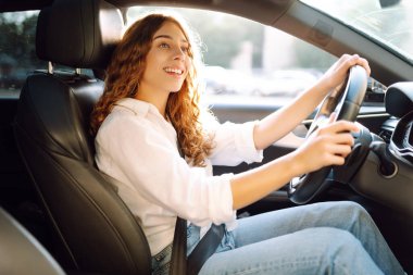 Kıvırcık saçlı genç bir kadın güneşin altında bir arabanın direksiyonunda oturur. Bir kadın sürücü güneşli bir günün tadını çıkarır. Taksi şöförü. Ulaşım kavramı, boş zaman.