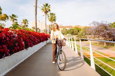 Bisikletli gülümseyen bir kadın şehirde güneşli bir günün tadını çıkarıyor. Güzel bir kadın parlak çiçeklerin arasında bisiklet sürüyor. Rahatlama konsepti, hafta sonu. Etkin yaşam biçimi.