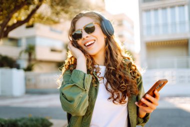 Kablosuz kulaklık takan, parkta yürürken müzik dinleyen genç bir kadının portresi. Güneş gözlüklü mutlu kadın akıllı telefon tutuyor ve güneşli günün tadını çıkarıyor. Yürüyüş, müzik ve açık hava kavramı.