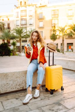 Şapkalı ve sarı valizli güzel bir turist güneşli meydanda telefonla konuşuyor. Telefonlu bir kadın şehirde mesajlaşıyor ve gezintiden zevk alıyor. Turizm, dinlenme, seyahat kavramı.