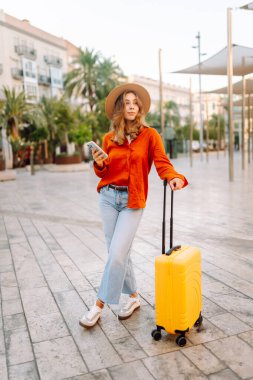 Şapkalı ve sarı valizli güzel bir turist güneşli meydanda telefonla konuşuyor. Telefonlu bir kadın şehirde mesajlaşıyor ve gezintiden zevk alıyor. Turizm, dinlenme, seyahat kavramı.