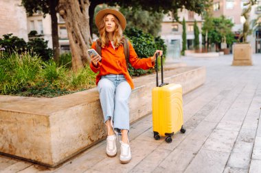 Şapkalı ve sarı valizli güzel bir turist güneşli meydanda telefonla konuşuyor. Telefonlu bir kadın şehirde mesajlaşıyor ve gezintiden zevk alıyor. Turizm, dinlenme, seyahat kavramı.