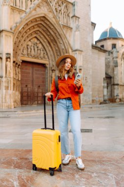 Şapkalı ve sarı valizli güzel bir turist güneşli meydanda telefonla konuşuyor. Telefonlu bir kadın şehirde mesajlaşıyor ve gezintiden zevk alıyor. Turizm, dinlenme, seyahat kavramı.