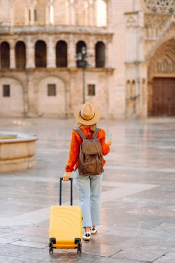 Şapkalı ve sarı valizli güzel bir turist güneşli meydanda telefonla konuşuyor. Telefonlu bir kadın şehirde mesajlaşıyor ve gezintiden zevk alıyor. Turizm, dinlenme, seyahat kavramı.