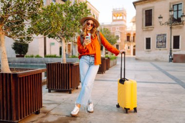 Şapkalı ve sarı valizli güzel bir turist güneşli meydanda telefonla konuşuyor. Telefonlu bir kadın şehirde mesajlaşıyor ve gezintiden zevk alıyor. Turizm, dinlenme, seyahat kavramı.