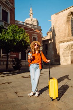 Şapkalı ve sarı valizli güzel bir turist güneşli meydanda telefonla konuşuyor. Telefonlu bir kadın şehirde mesajlaşıyor ve gezintiden zevk alıyor. Turizm, dinlenme, seyahat kavramı.