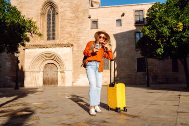 Şapkalı ve sarı valizli güzel bir turist güneşli meydanda telefonla konuşuyor. Telefonlu bir kadın şehirde mesajlaşıyor ve gezintiden zevk alıyor. Turizm, dinlenme, seyahat kavramı.