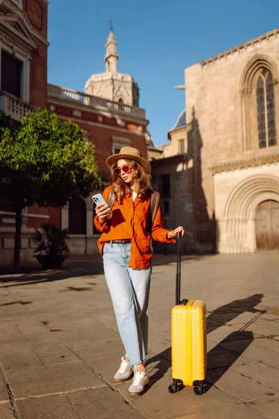 Şapkalı ve sarı valizli güzel bir turist güneşli meydanda telefonla konuşuyor. Telefonlu bir kadın şehirde mesajlaşıyor ve gezintiden zevk alıyor. Turizm, dinlenme, seyahat kavramı.