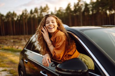 Gülümseyen bir kadın arabanın camından sarkar ve günbatımının tadını çıkarır. Araba macerasına çıkmış güzel bir kadın güneş ışınlarında kendini özgür hisseder. Özgürlük, macera ve araba kavramı..