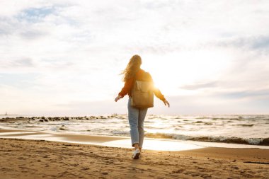 Sırt çantalı biri güneşli bir sahilde kendini özgür hisseder. Genç bir kadın deniz kıyısında geziniyor ve gün batımında doğanın tadını çıkarıyor. Yürüme, rahatlama ve deniz kavramı.