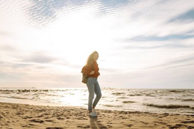 Sırt çantalı biri güneşli bir sahilde kendini özgür hisseder. Genç bir kadın deniz kıyısında geziniyor ve gün batımında doğanın tadını çıkarıyor. Yürüme, rahatlama ve deniz kavramı.