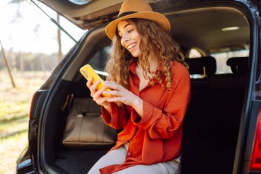 Telefonu olan neşeli bir kadın bir araba yolculuğunda arabanın bagajında oturur. Güzel bir kadın güneşli bir günde doğanın tadını çıkarır. Macera kavramı, hafta sonu.