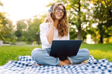 Sunny Park 'ta elinde dizüstü bilgisayarla battaniyenin üzerinde oturan serbest çalışan bir kadın. Kablosuz kulaklık takan kadın laptopta çalışıyor ve açık havada güneşli bir günün tadını çıkarıyor. Kavram, teknoloji, doğa ve rahatlama..
