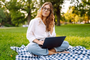 Sunny Park 'ta elinde dizüstü bilgisayarla battaniyenin üzerinde oturan serbest çalışan bir kadın. Kablosuz kulaklık takan kadın laptopta çalışıyor ve açık havada güneşli bir günün tadını çıkarıyor. Kavram, teknoloji, doğa ve rahatlama..