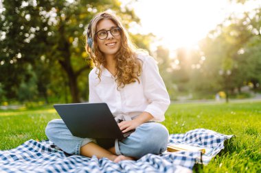 Sunny Park 'ta elinde dizüstü bilgisayarla battaniyenin üzerinde oturan serbest çalışan bir kadın. Kablosuz kulaklık takan kadın laptopta çalışıyor ve açık havada güneşli bir günün tadını çıkarıyor. Kavram, teknoloji, doğa ve rahatlama..