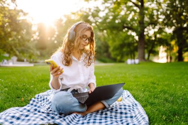 Sunny Park 'ta elinde dizüstü bilgisayarla battaniyenin üzerinde oturan serbest çalışan bir kadın. Kablosuz kulaklık takan kadın laptopta çalışıyor ve açık havada güneşli bir günün tadını çıkarıyor. Kavram, teknoloji, doğa ve rahatlama..