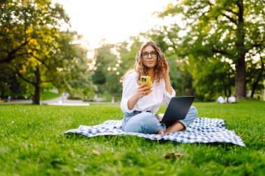 Sunny Park 'ta elinde dizüstü bilgisayarla battaniyenin üzerinde oturan serbest çalışan bir kadın. Kablosuz kulaklık takan kadın laptopta çalışıyor ve açık havada güneşli bir günün tadını çıkarıyor. Kavram, teknoloji, doğa ve rahatlama..